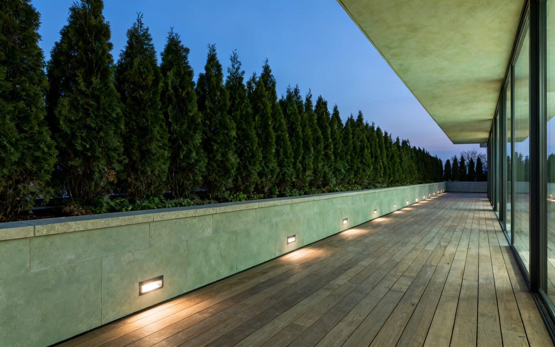 A long terrace at dusk under a deep blue sky. A tall conifer hedge runs behind a limestone planter wall, recessed floor lights wash the oak decking, and a glazed wall reflects the last light on the right.