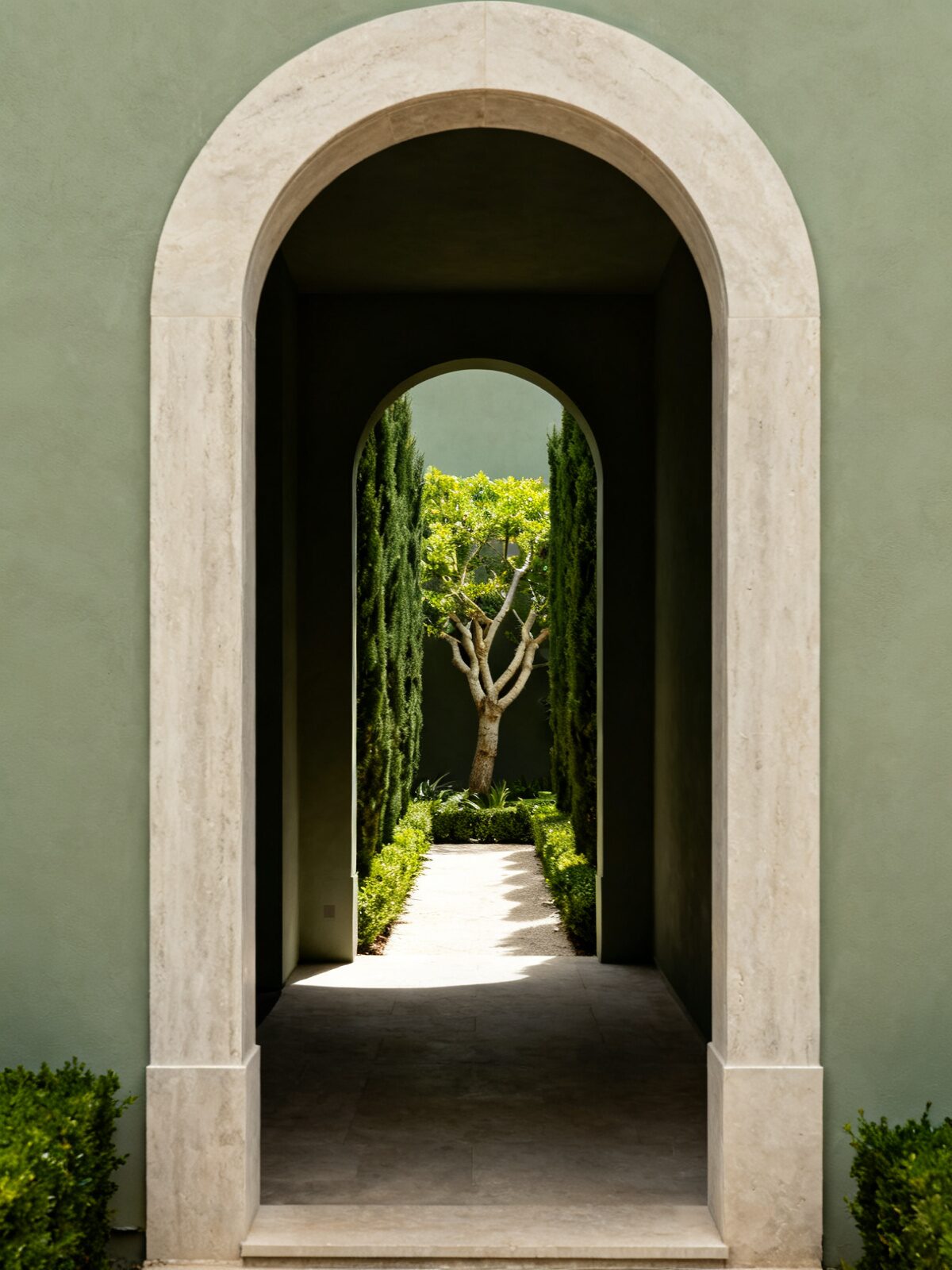 A tall honed limestone arch opening into a dark passage, with a second arch beyond it framing a narrow courtyard of clipped cypress and a single multi-stem tree.