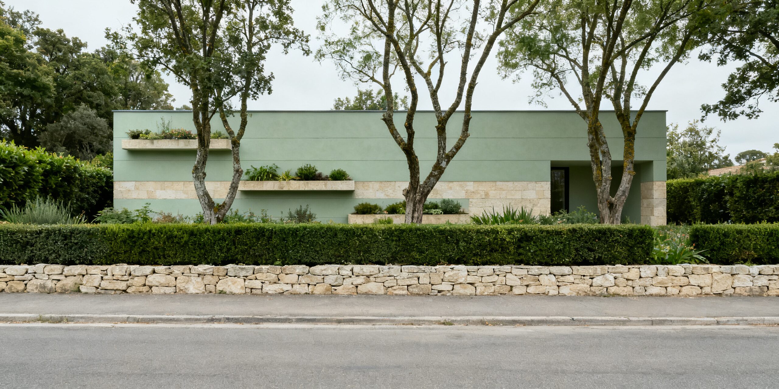 A low building in pale green plaster set back behind a dry stone boundary wall and a clipped hedge, with mature trees rising in front of it and an empty road across the foreground.