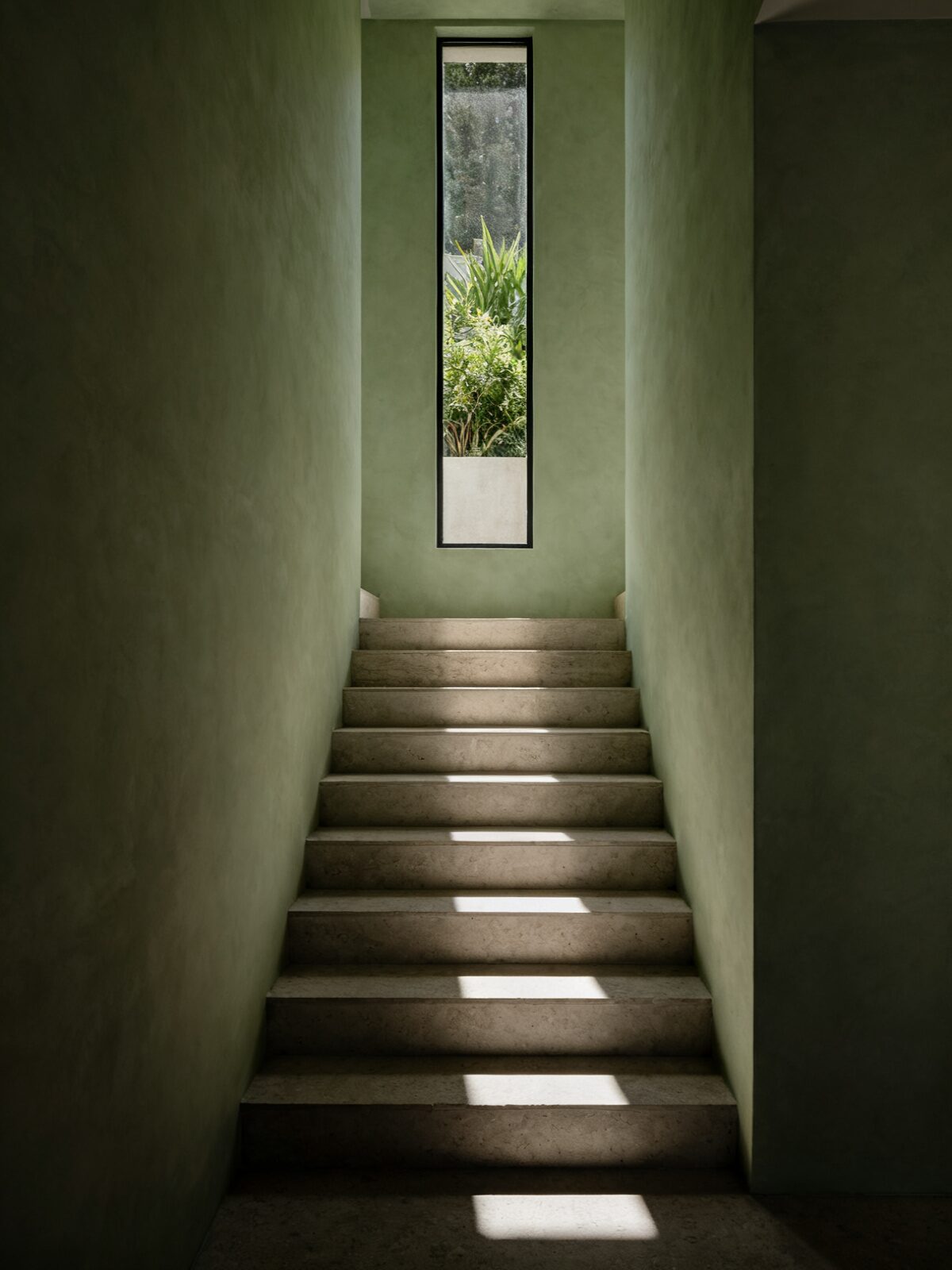 A limestone stair rising between bare lime plaster walls toward a tall narrow slot window, with terrace planting visible through the glass and one shaft of low sun falling across the treads.
