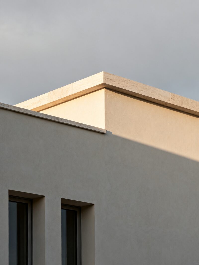 The top corner of a pale plaster building, where a honed limestone coping catches low warm sun against an overcast grey sky, with two deep window reveals cut into the wall below.